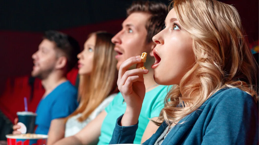 a row of people sitting in a movie theatre eating popcorn and looking in awe at a promo video
